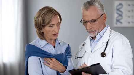 Un médico con estetoscopio y bata toma notas mientras una paciente se queda mirando.