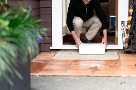 An older man stoops down to pick up a package sitting on his doorstep