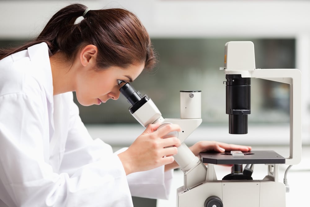 Una mujer con bata de laboratorio mirando por un microscopio.