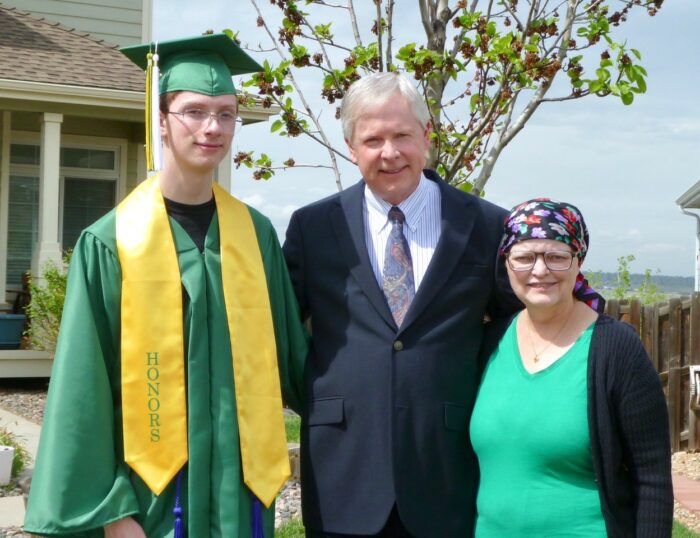 A young man dressed in a cap and gown for his graduation poses with his parents. His mother wears a head scarf.