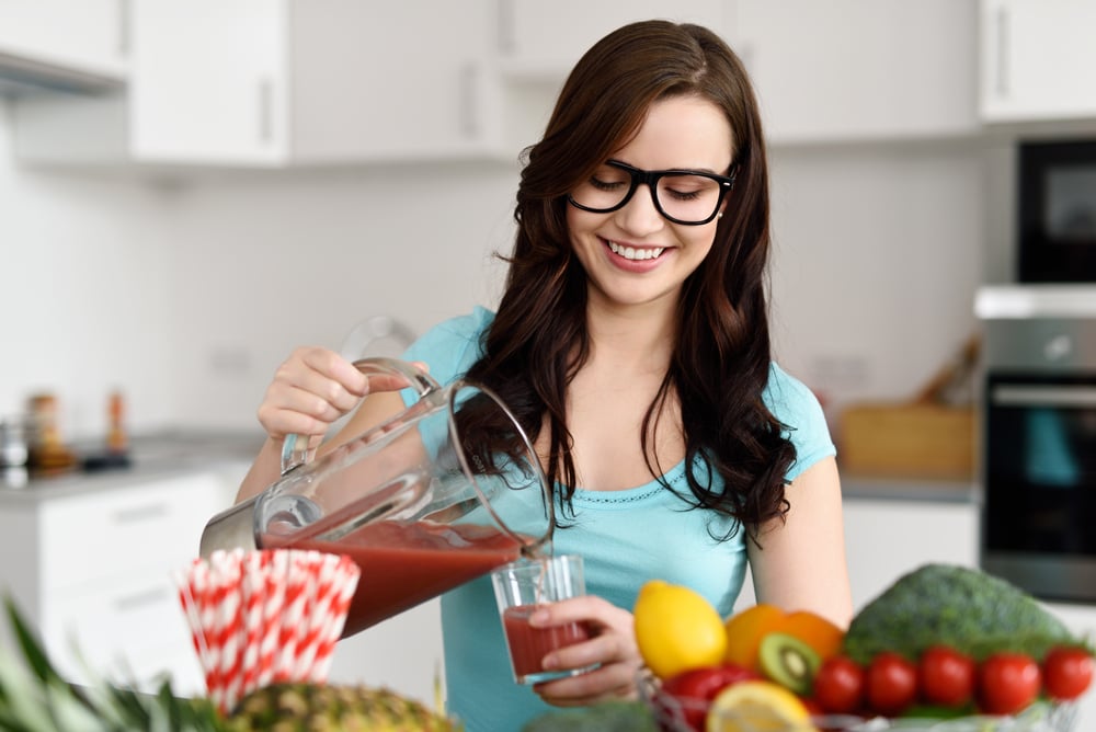 Joven feliz y saludable con gafas sirviendo batidos de verduras recién hechos con ingredientes vegetales variados en la encimera de su cocina