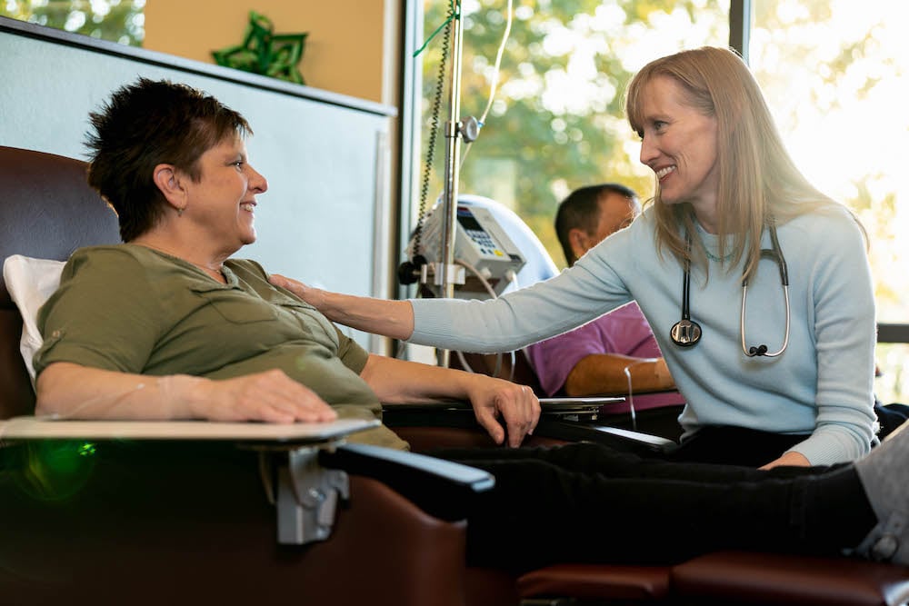 A medical provider wearing a stethoscope comforts a woman sitting in a chair by smiling and putting her hand on her shoulder.